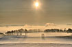 The sun is shining through the morning mist covering trees and snow covered fields