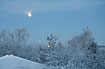 The full moon descends behind snow covered trees and roofs